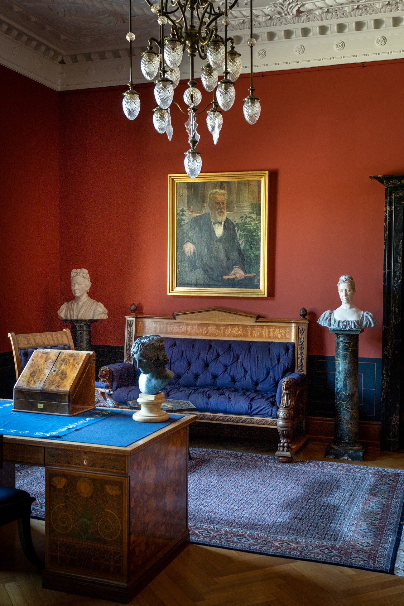 Elegant interior room with red walls, crystal chandelier, blue upholstered sofa, marble busts, and framed portrait