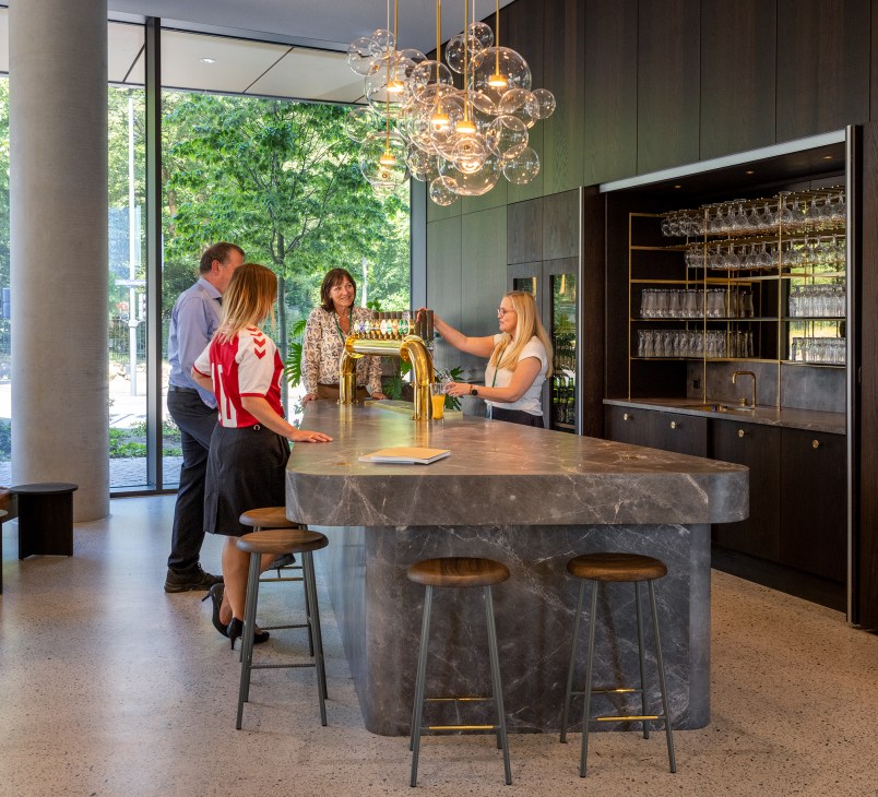 Modern office bar area with marble island, brass tap, glass storage, and employees gathered around