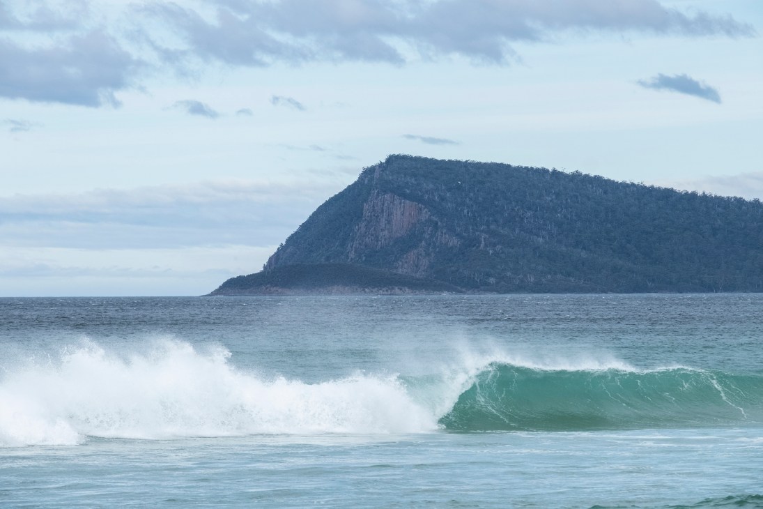 Ocean waves crashing onto beach with forested headland in background under cloudy sky