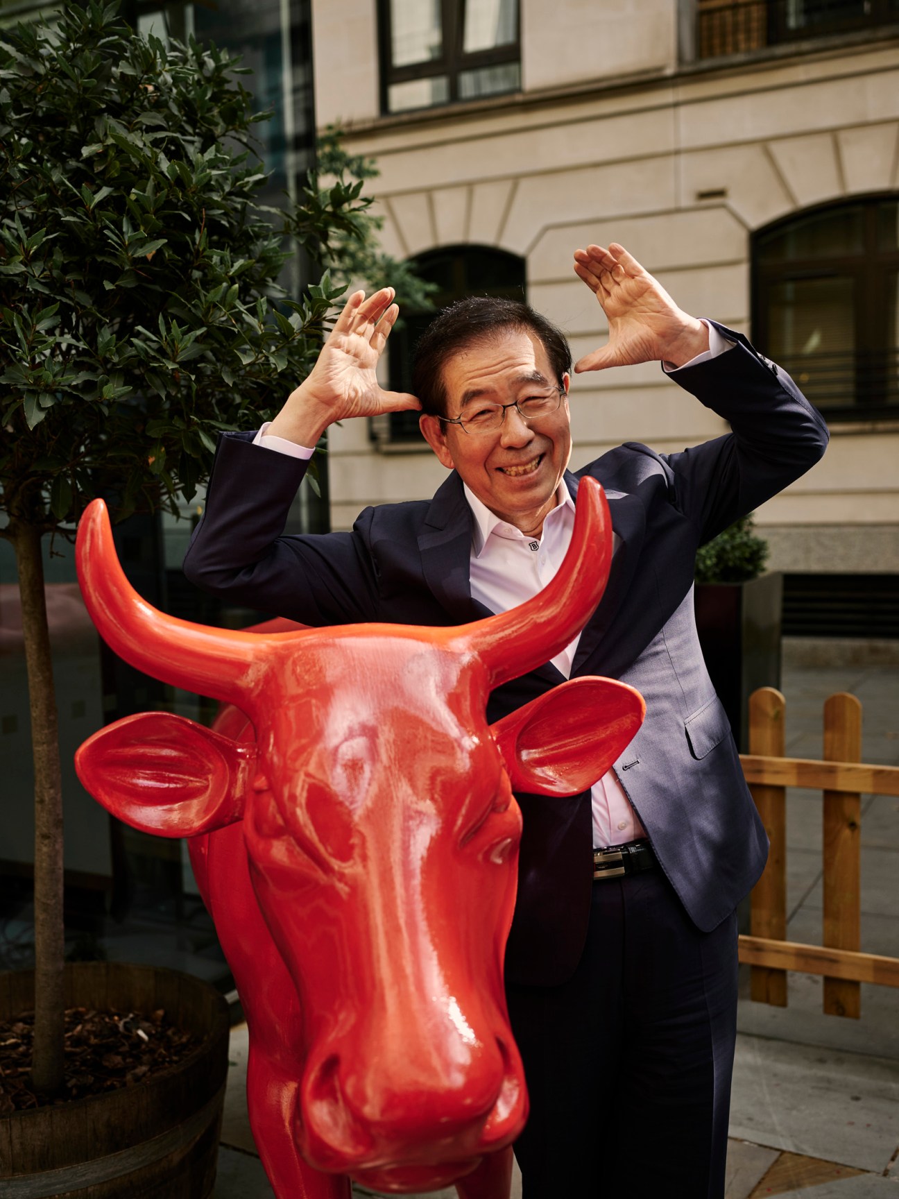 Park Won-soon, mayor of Seoul, poses playfully with a red bull sculpture outdoors.
