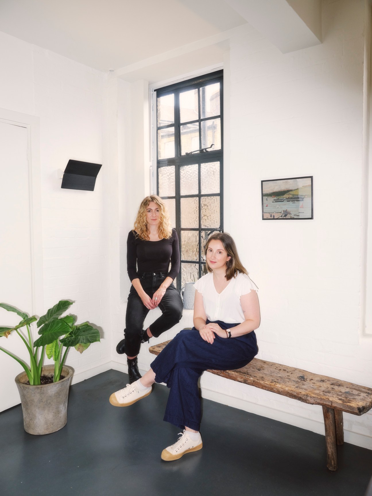 Two women sitting in modern minimalist office space with white walls, wooden bench, and large gridded window