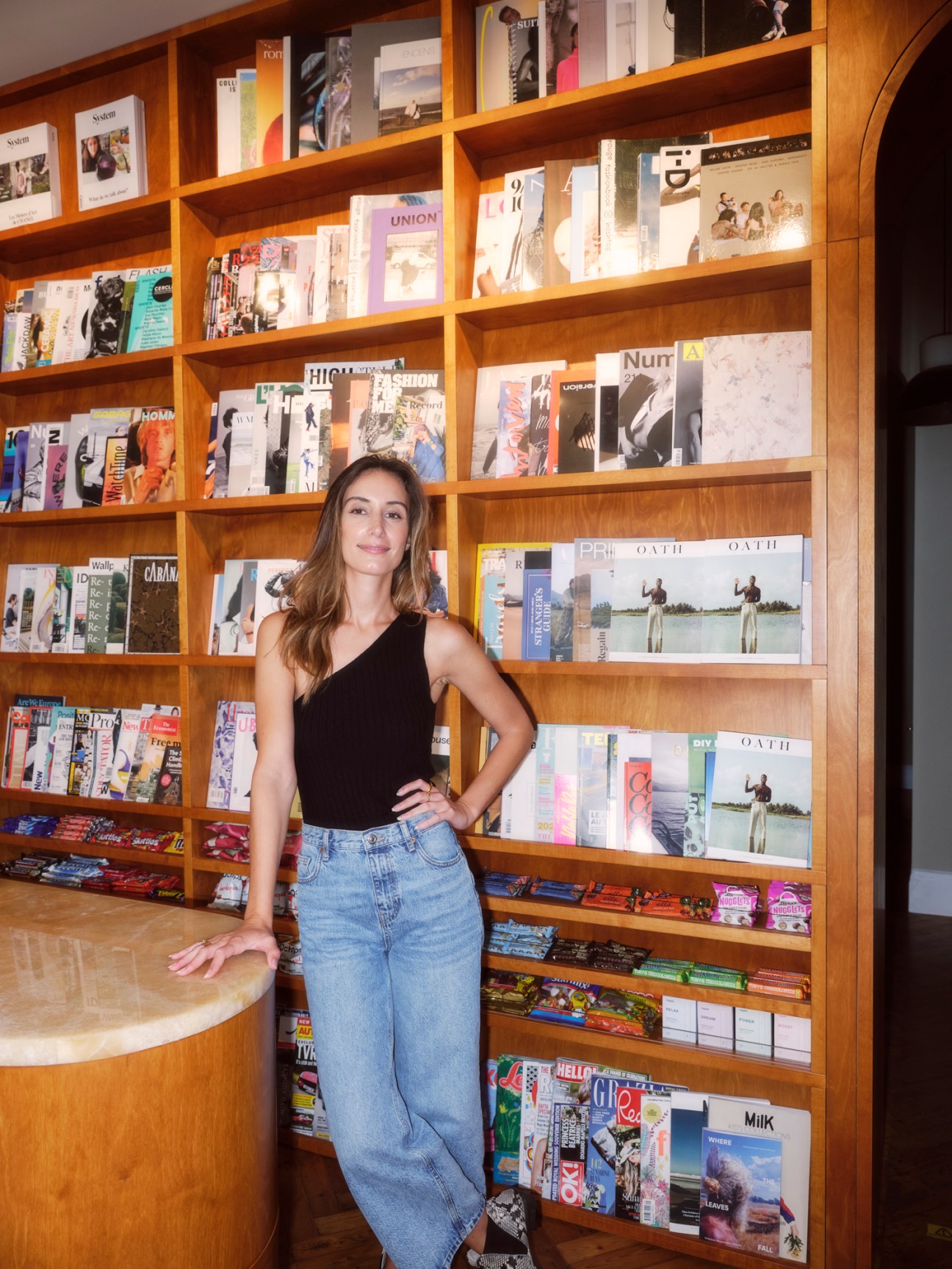 Stephanie Blomkamp of 'Oath' magazine standing in front of wooden magazine display shelves