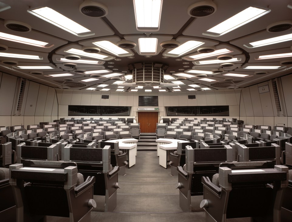 Interior of Saal 6 conference room with rows of black and gray delegate seating, geometric ceiling panels with recessed lighting, and an exit door at the far end.