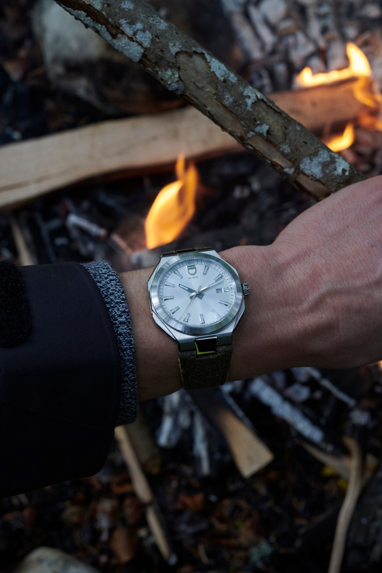 A wrist wearing an ID Genève watch with white dial and dark strap, photographed near a campfire