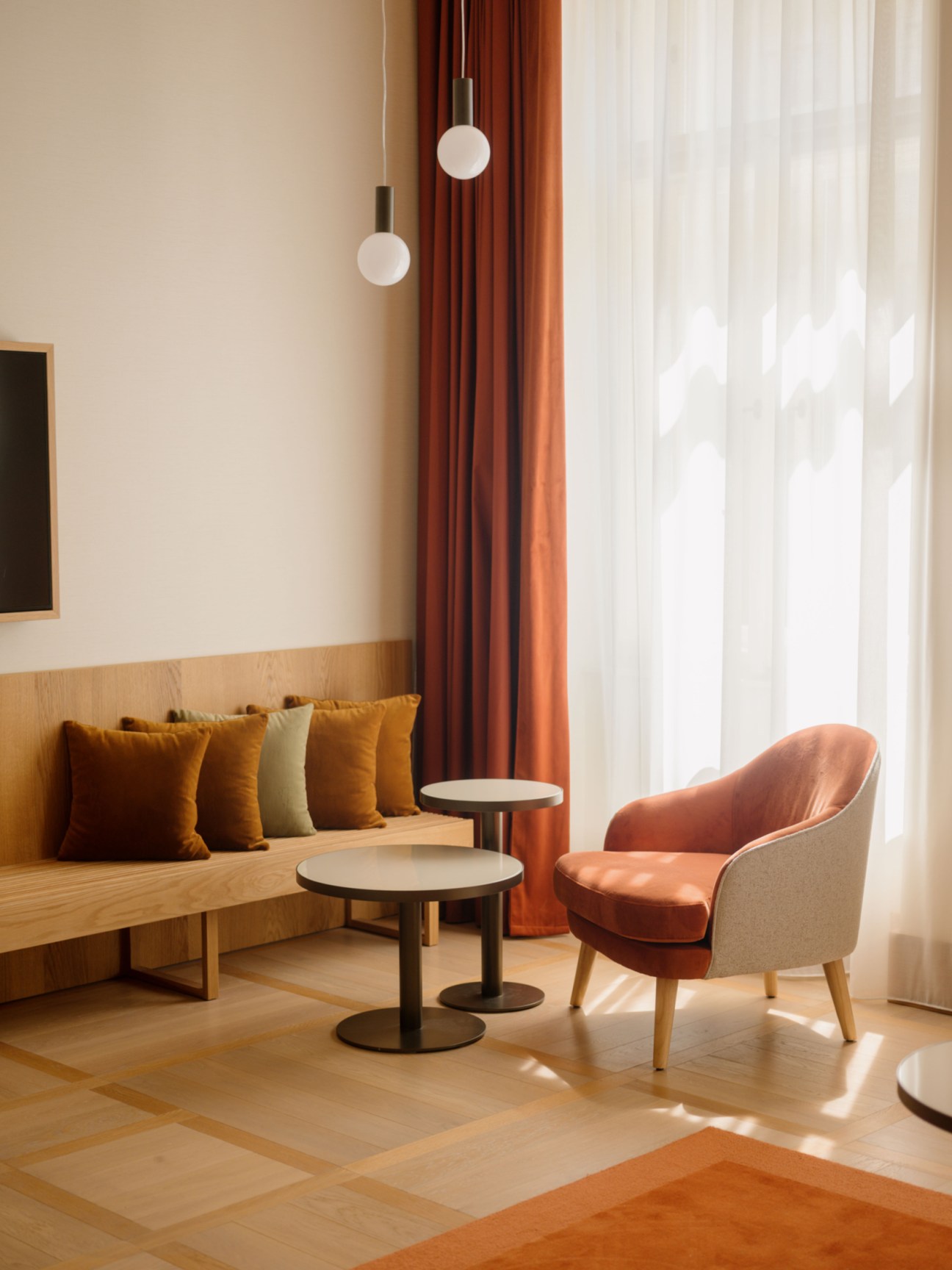 Modern hotel seating area with coral chair, wooden bench with orange pillows, and pendant lights by window