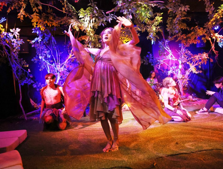 Performer in flowing dress dances center stage in forest-themed theatrical set with colorful lighting and other actors