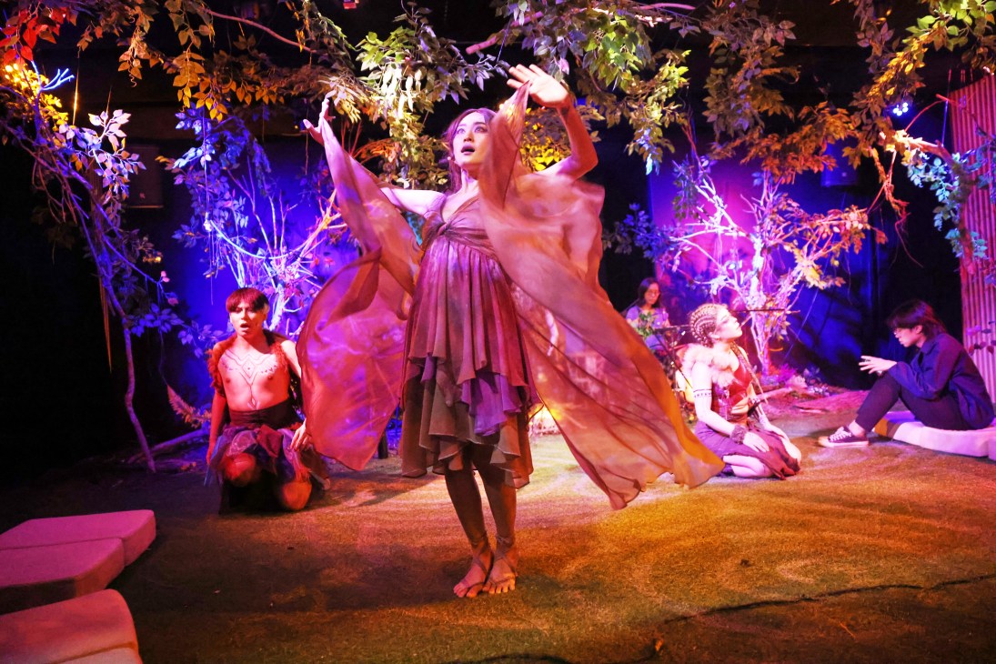 Performer in flowing dress with raised arms on intimate theater stage with purple lighting and foliage backdrop