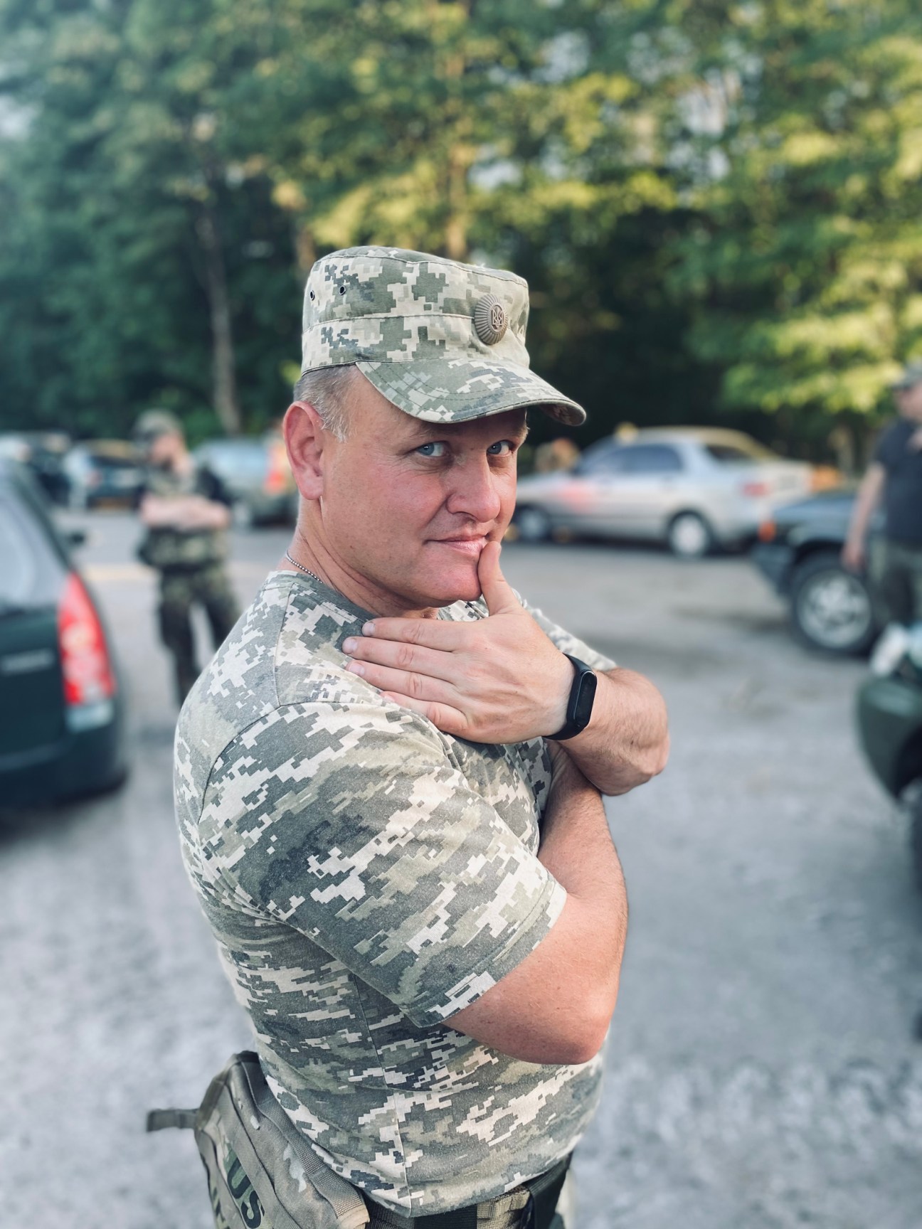 Ukrainian military reservist in digital camouflage uniform and cap standing in a parking lot with trees in background.