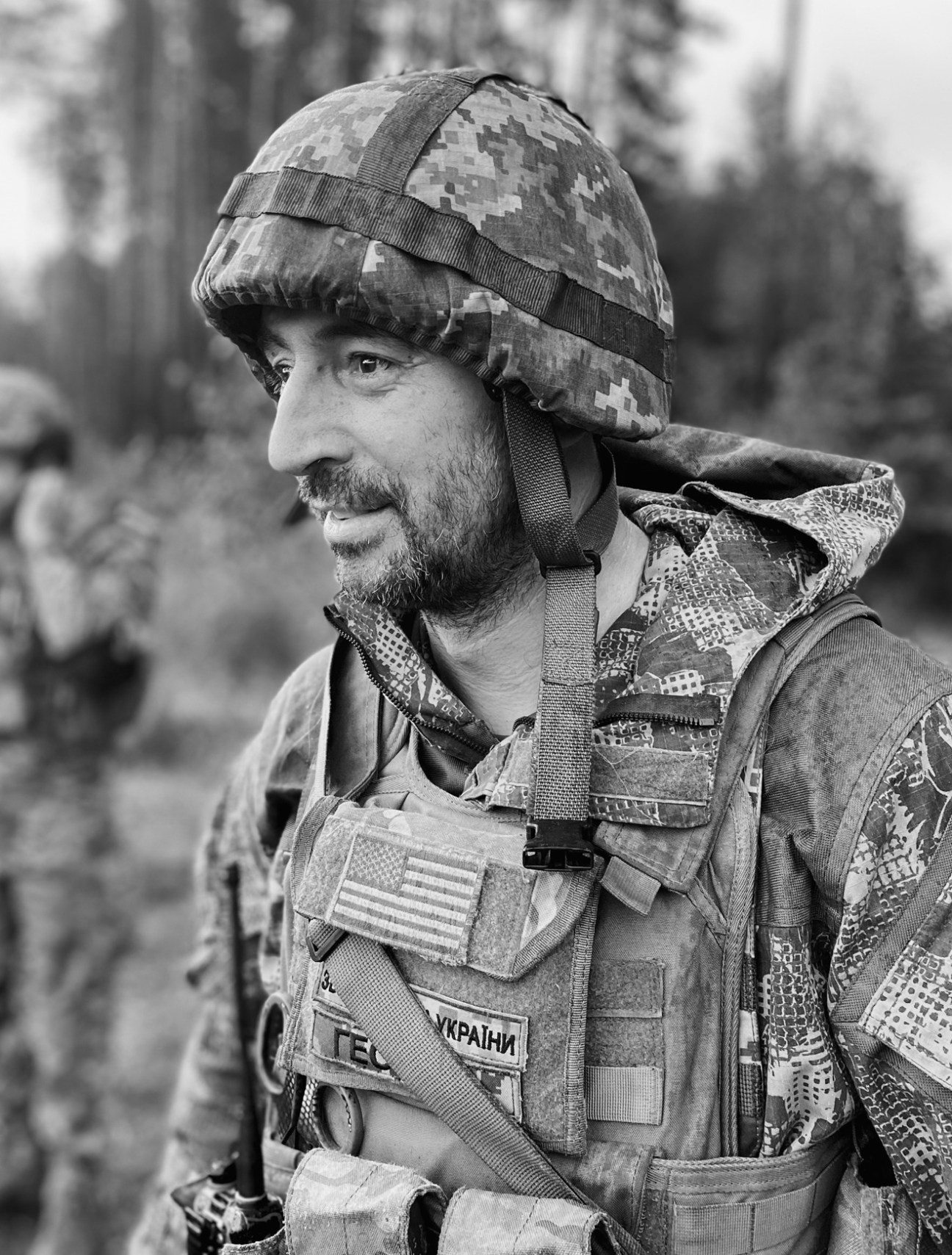 Ukrainian soldier in camouflage helmet and military fatigues with Ukrainian flag patch, photographed in profile in a wooded area.