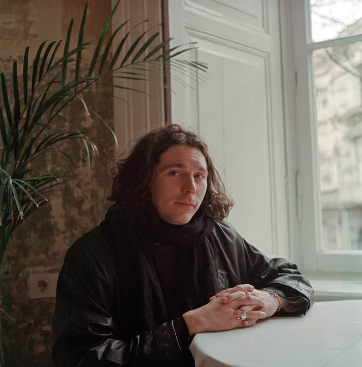 Denis Klimov, a young man with curly hair wearing a dark jacket, sits at a white table by a window in Tbilisi