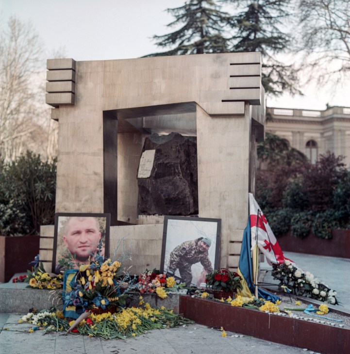 Memorial monument with concrete frame housing dark stone, surrounded by portrait photos, flowers and Ukrainian flag