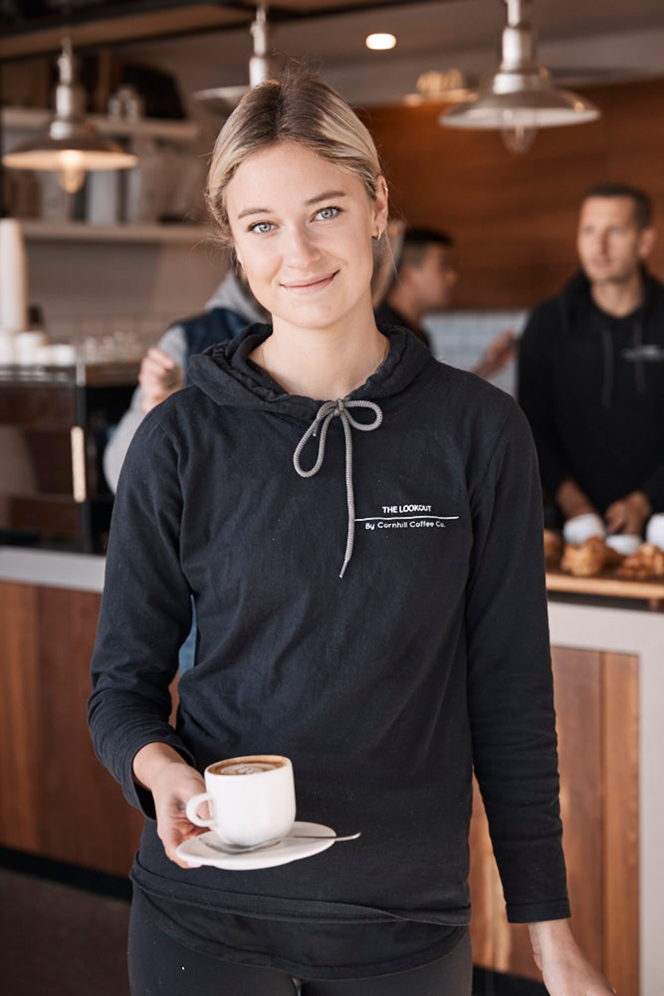 Blonde barista in black hoodie holds white coffee cup in modern café with industrial lighting and customers in background
