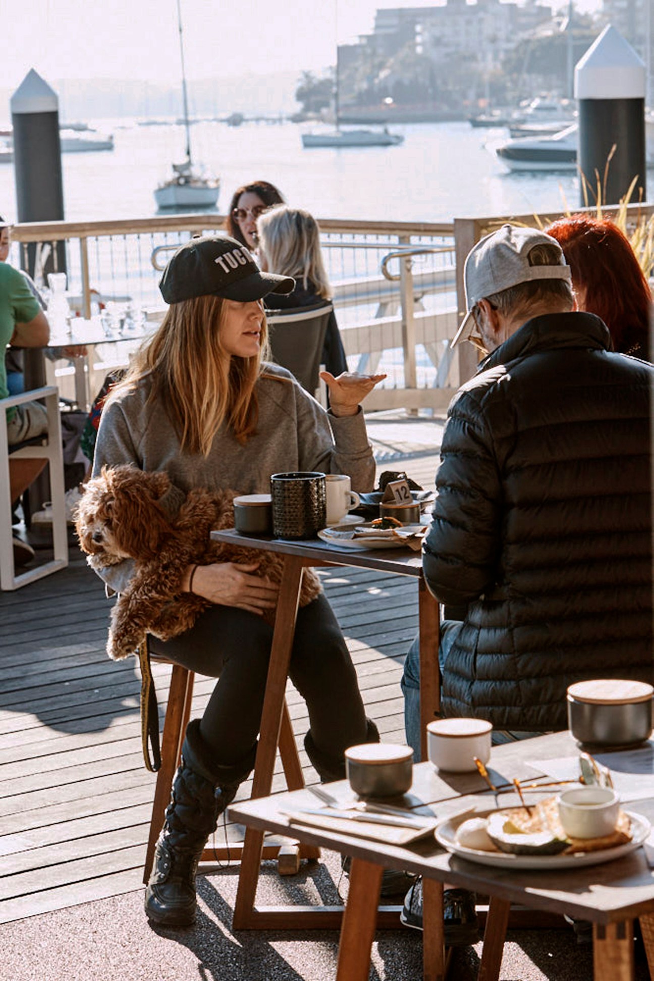 Woman in cap holding small dog at waterfront cafe with boats in harbor background