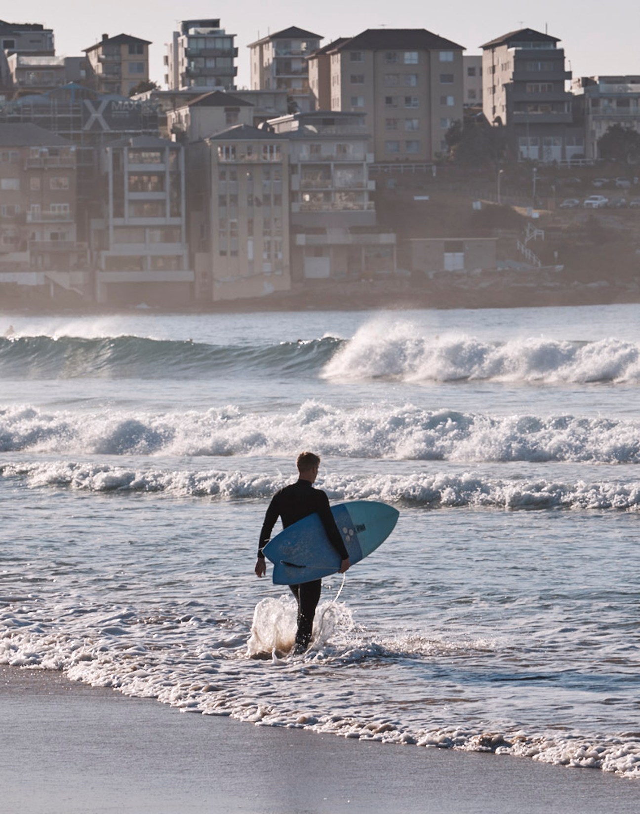 A surfer in wetsuit carrying a blue surfboard walks into ocean waves at a beach
