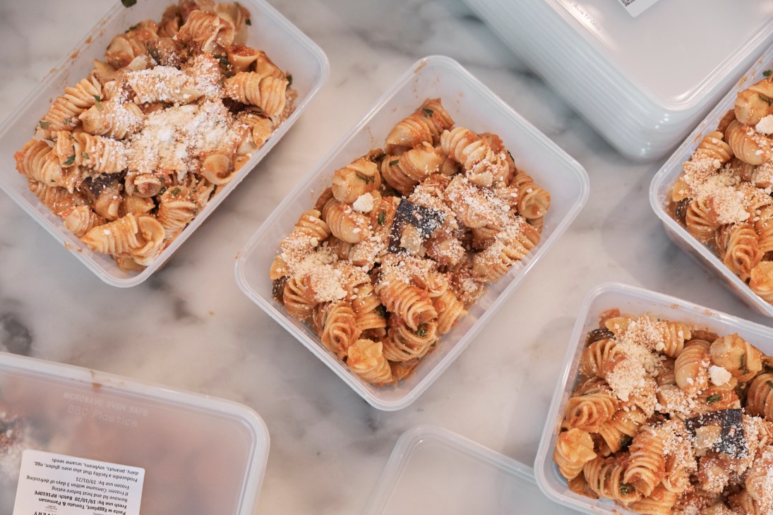 Plastic containers of pasta with ragù sauce topped with grated cheese on a marble surface.