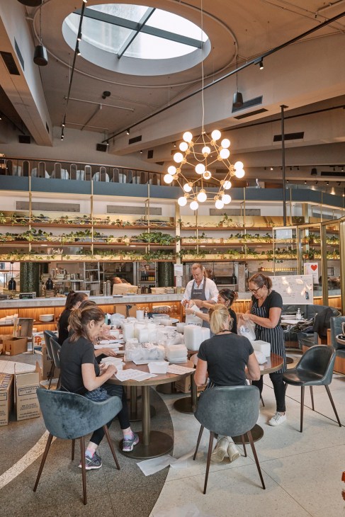 Volunteers pack meals at a table in a modern kitchen with skylights and pendant lighting.