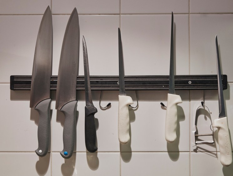 Various kitchen knives with different blade sizes and handle colors mounted on a black magnetic strip against white tile wall.
