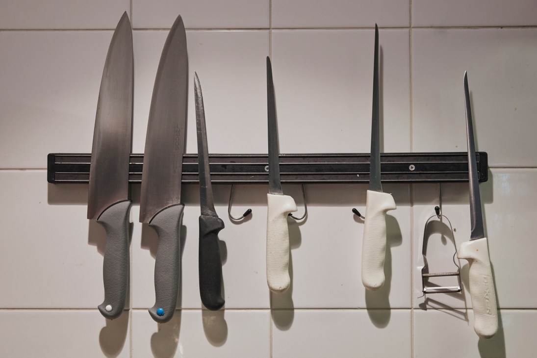 Various kitchen knives with different blade sizes and handle colors mounted on a black magnetic strip against white tile wall.