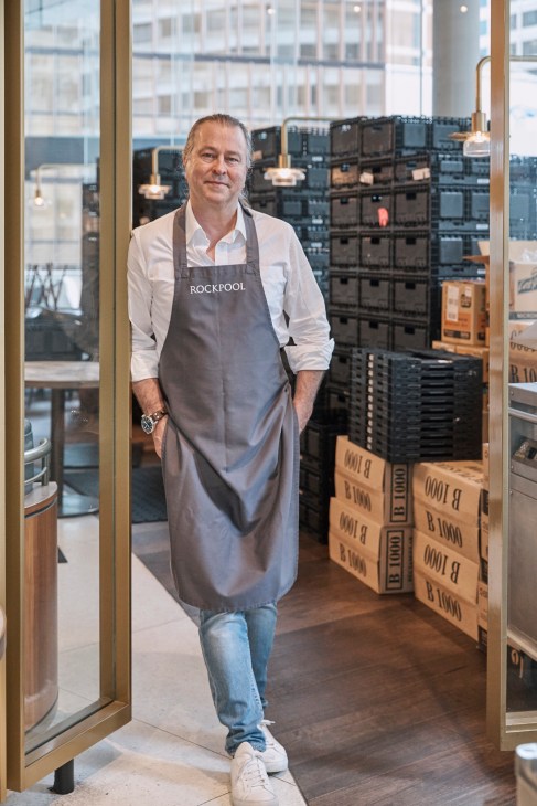Chef Neil Perry wearing a Rockpool apron stands at Hope Delivery headquarters with stacked crates behind him.