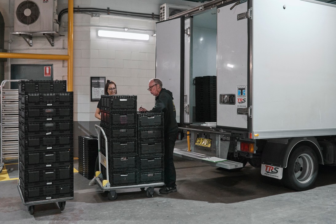Two people load black plastic crates of meals onto an OzHarvest delivery truck in a warehouse.