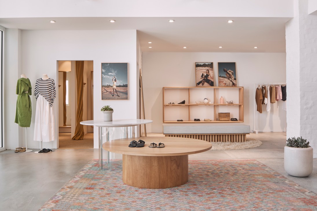 Interior of a minimalist clothing retail shop with wooden display tables, neutral-toned garments on rails, and natural lighting.