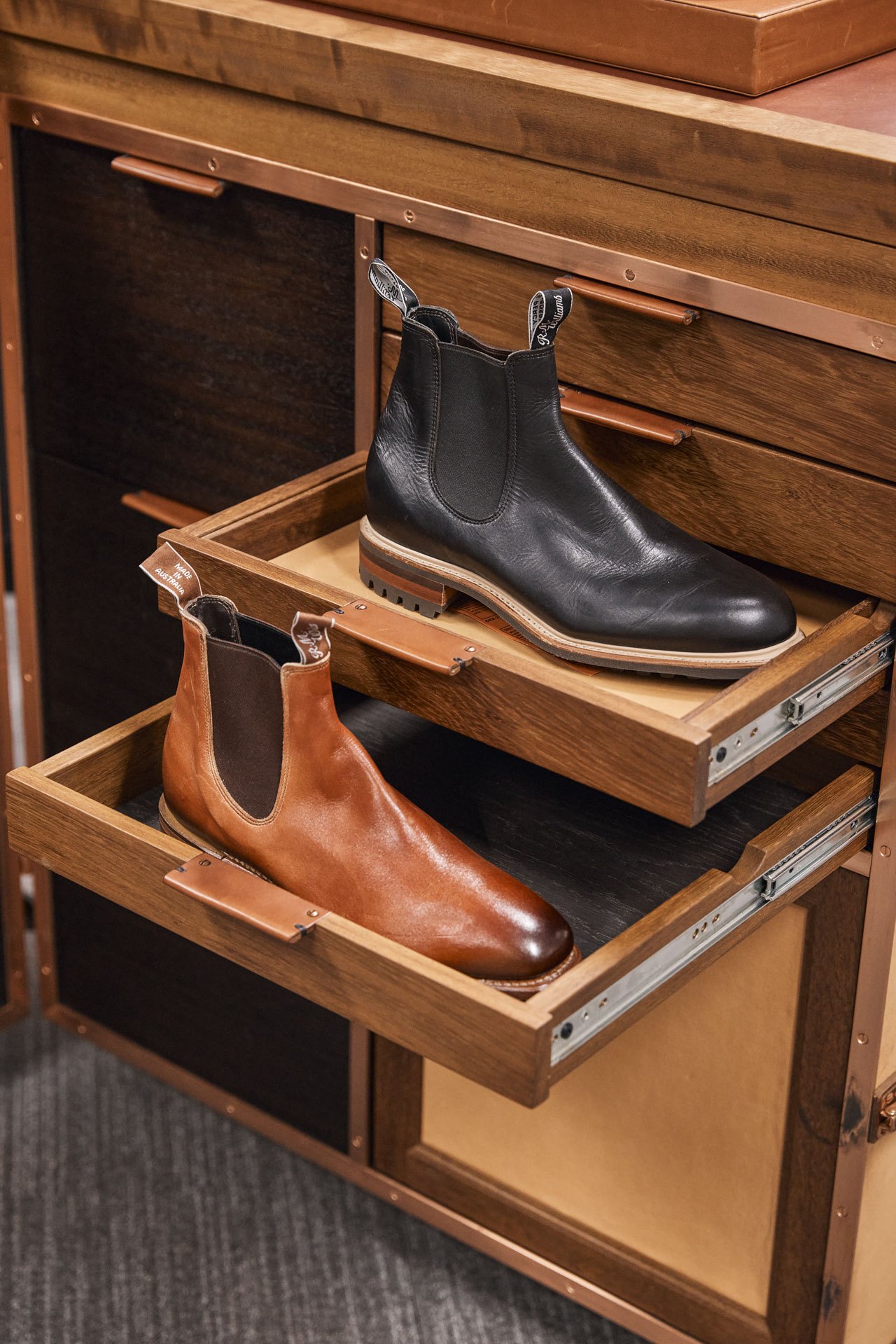 Two RM Williams leather boots displayed on pull-out wooden drawers in an organized storage cabinet