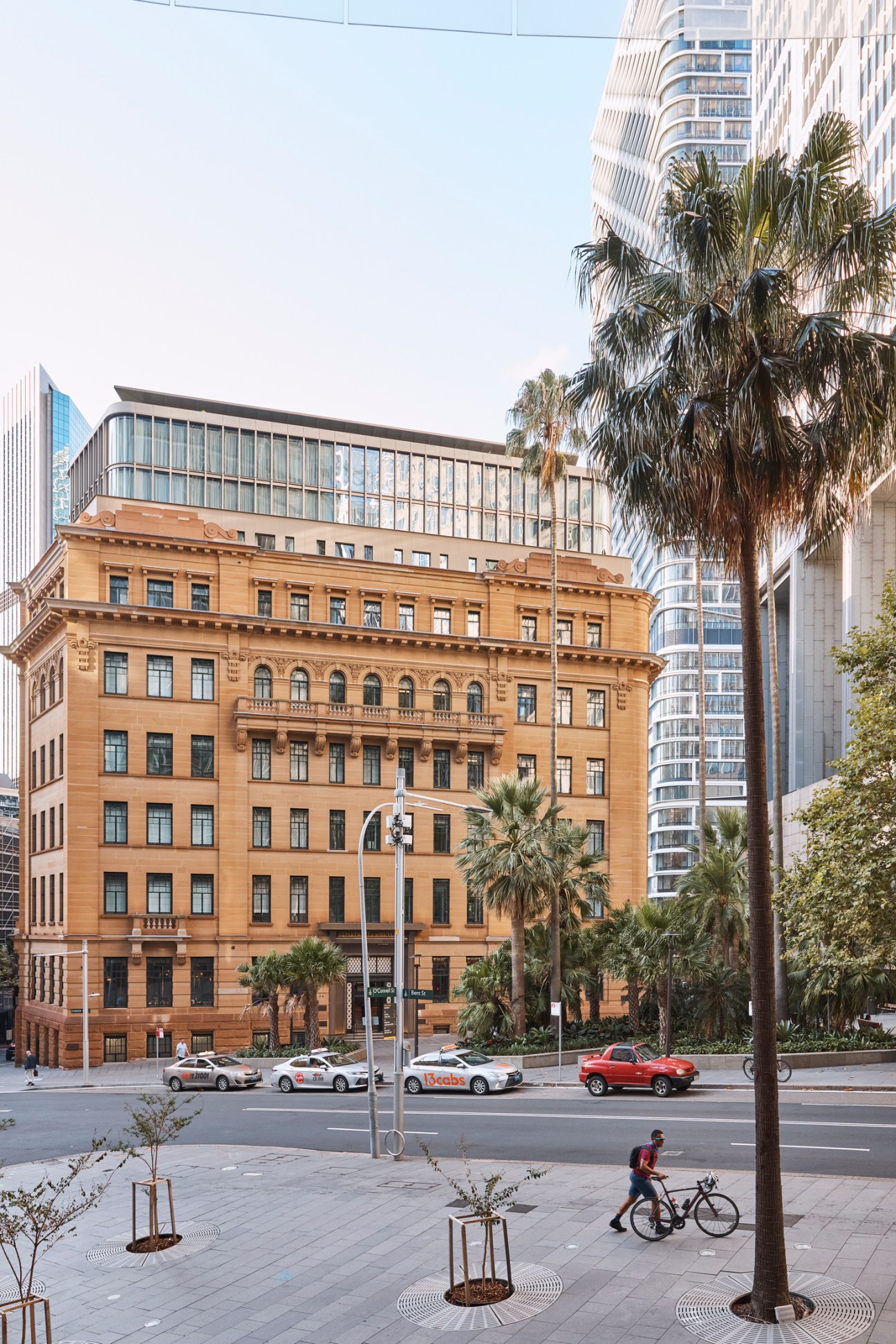 Historic sandstone building with classical architecture, modern glass addition on top, surrounded by palm trees and skyscrapers