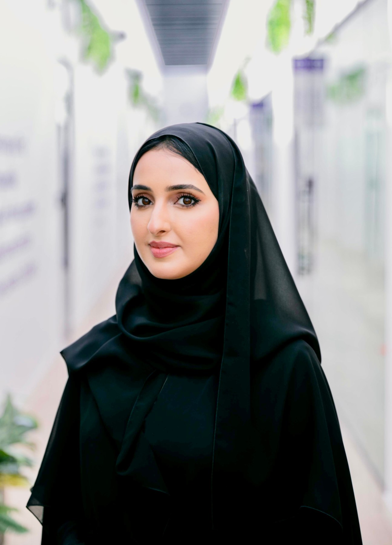 Portrait of Ebtesam Almazrouei, a woman wearing a black hijab, standing in a modern corridor with green plants and white walls in the background.