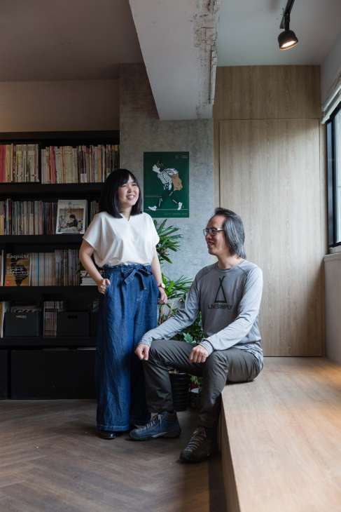 Two people pose in a modern office space with bookshelves and plants, one standing and one crouching.