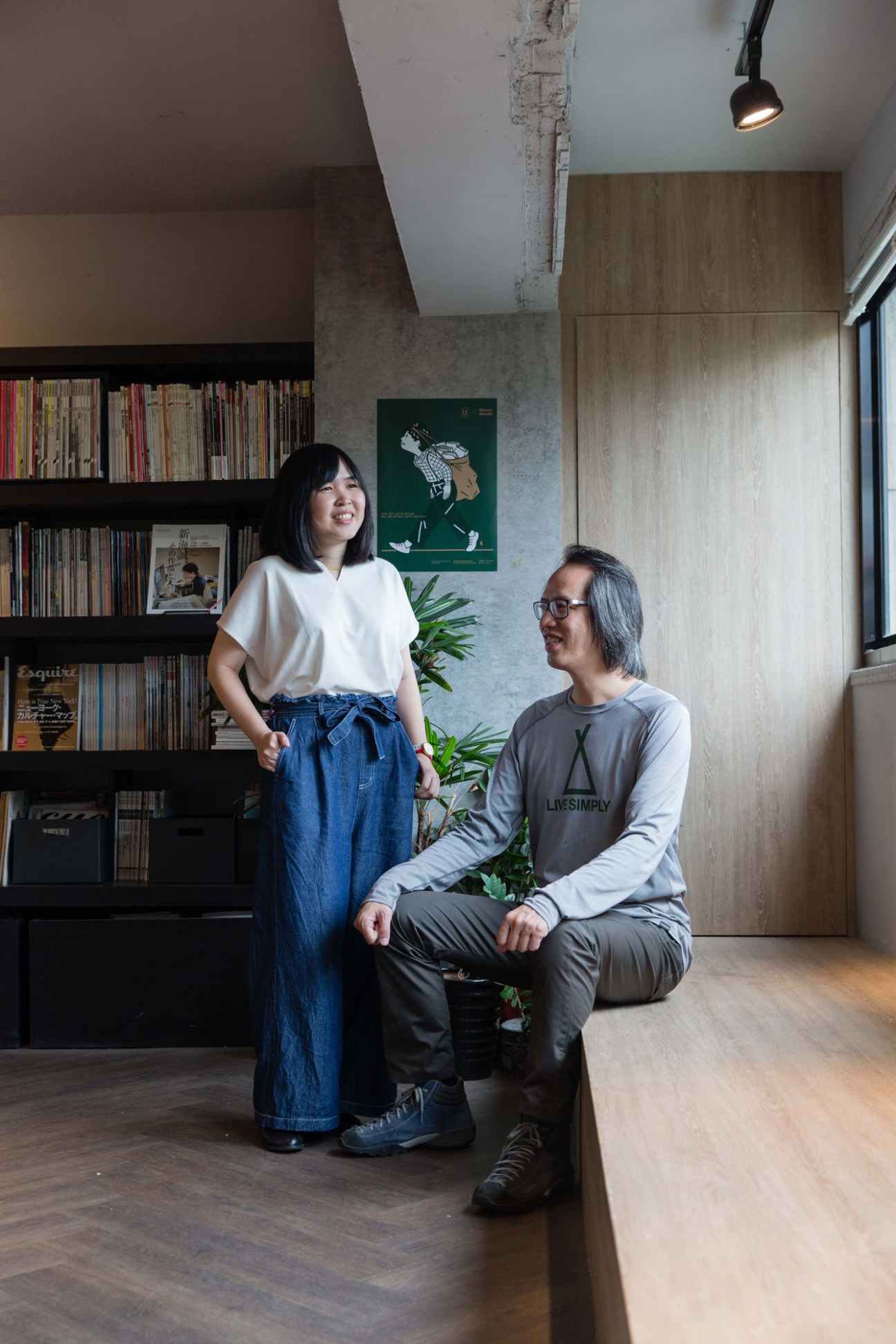Two people in a modern office space with bookshelves, one standing and one crouching, both smiling at each other.