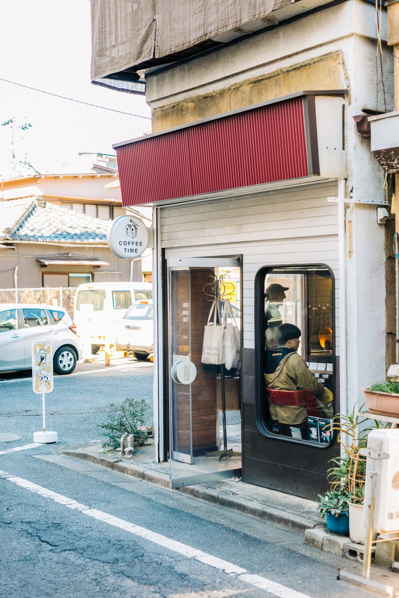 Small coffee shop storefront with red awning, person visible inside, white car parked on street