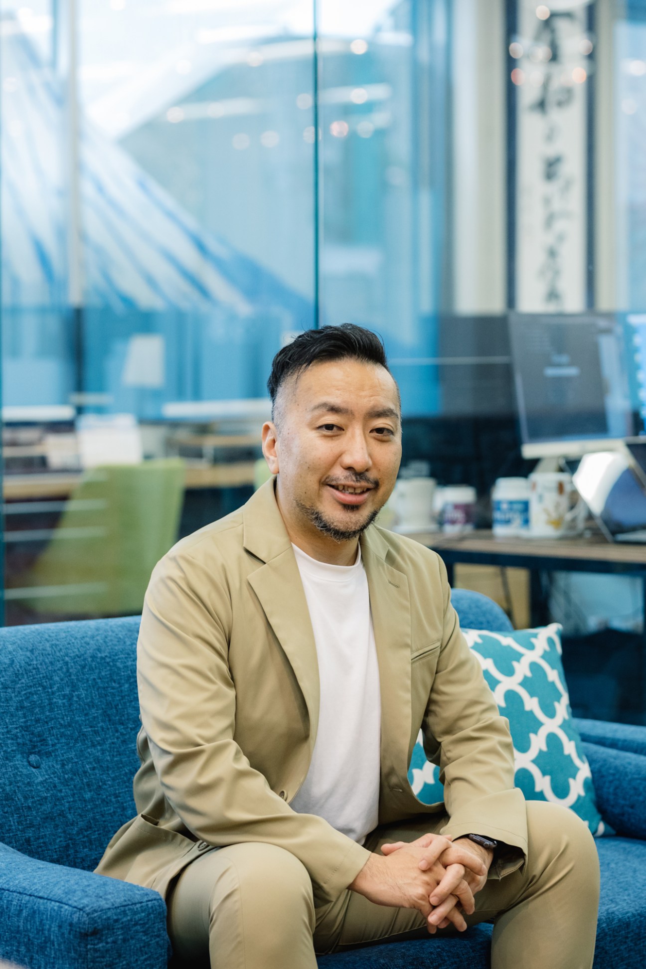 Man in beige blazer and white shirt smiling in modern office with blue sofa and city views