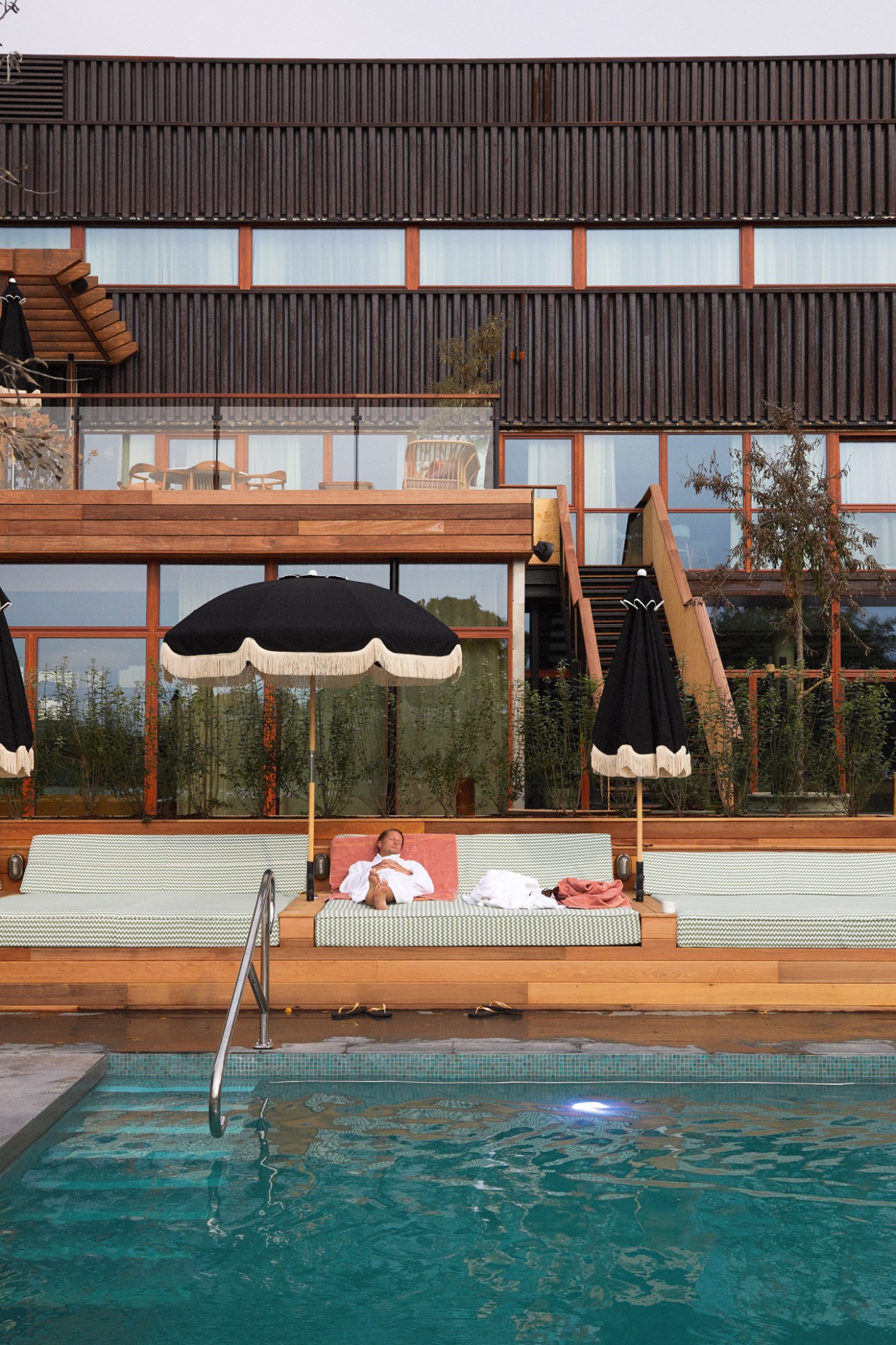 Person relaxing on wooden deck lounger by heated outdoor pool at Ellery Beach House Stockholm