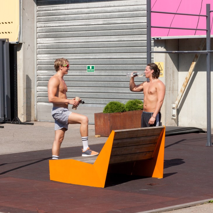 Two shirtless men exercising outdoors with dumbbells near a modern orange and wood bench against an industrial backdrop.