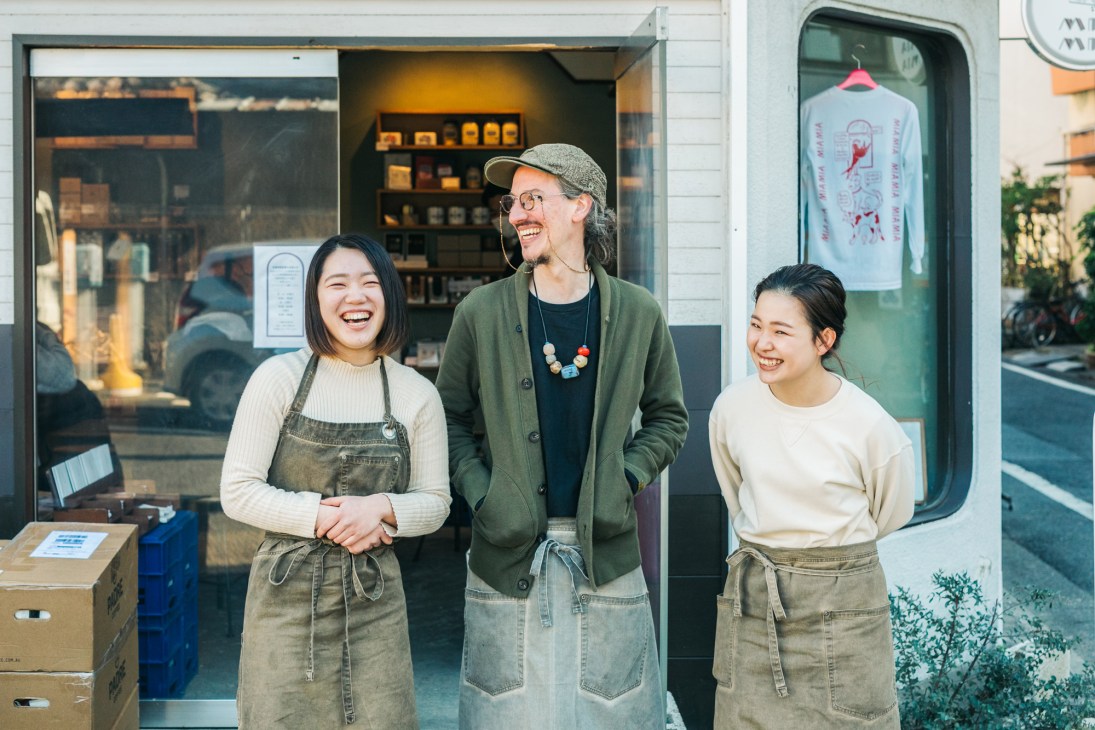 Three people in aprons standing together and smiling outside a cafe storefront
