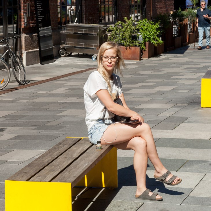Woman sitting on a yellow and wood Vestre Bloc bench in a sunny urban plaza with brick buildings and storefronts.