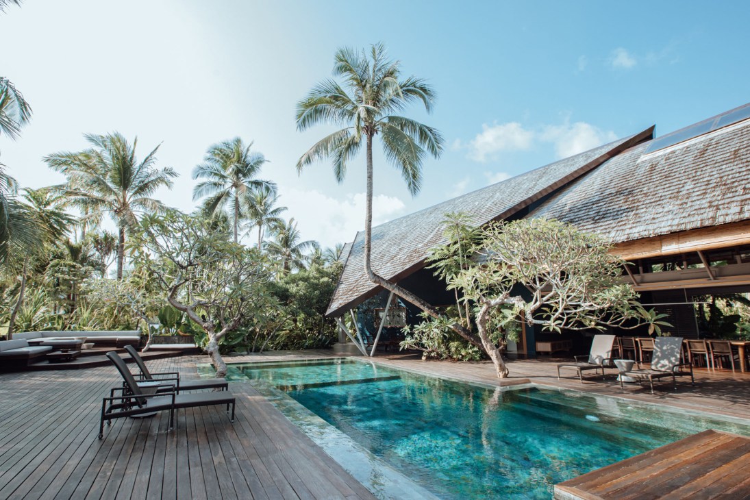 Swimming pool surrounded by wooden decking with tropical palm trees and traditional roofed buildings