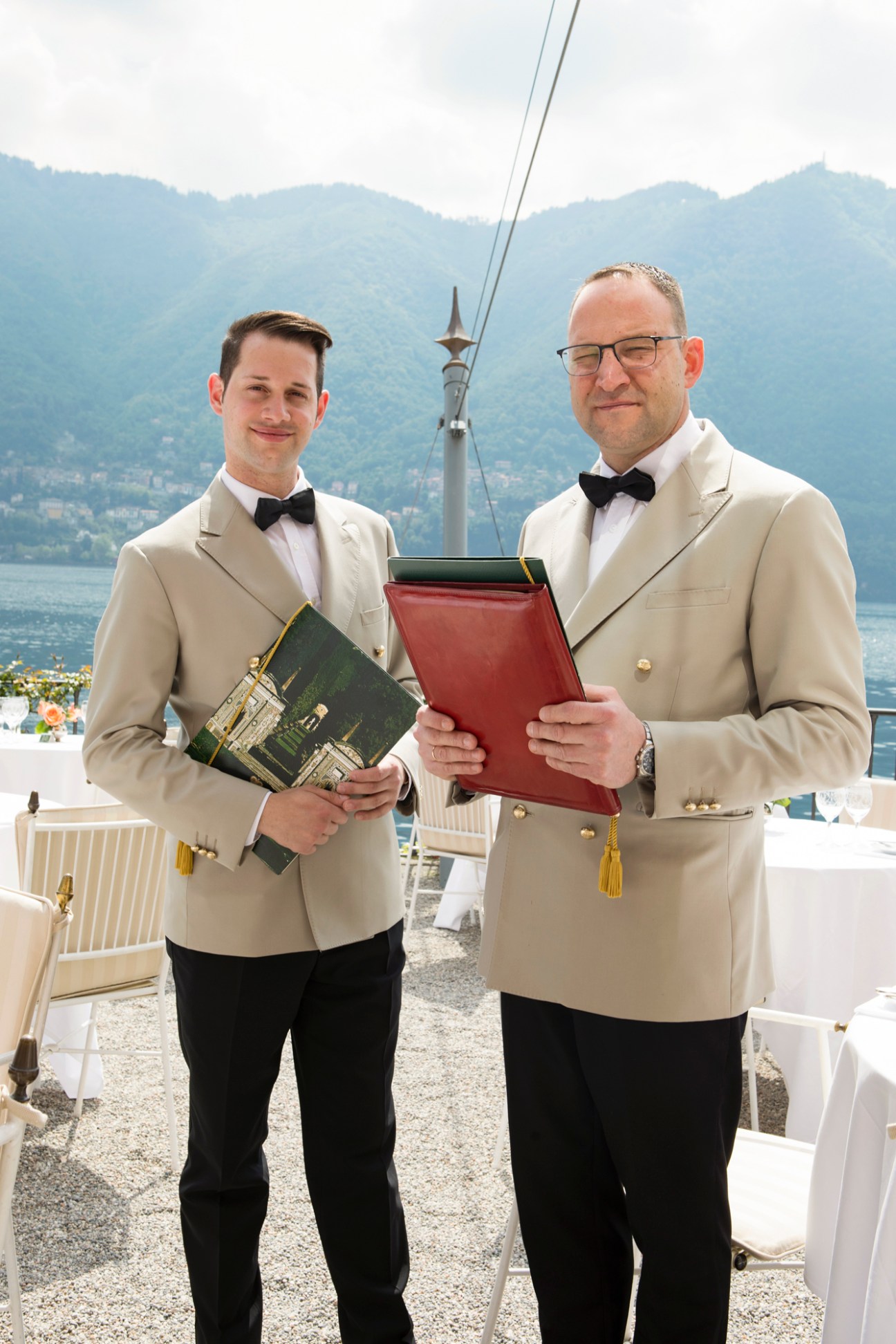 Two hotel staff members in cream blazers and bow ties holding menus on outdoor terrace with lake and mountains behind