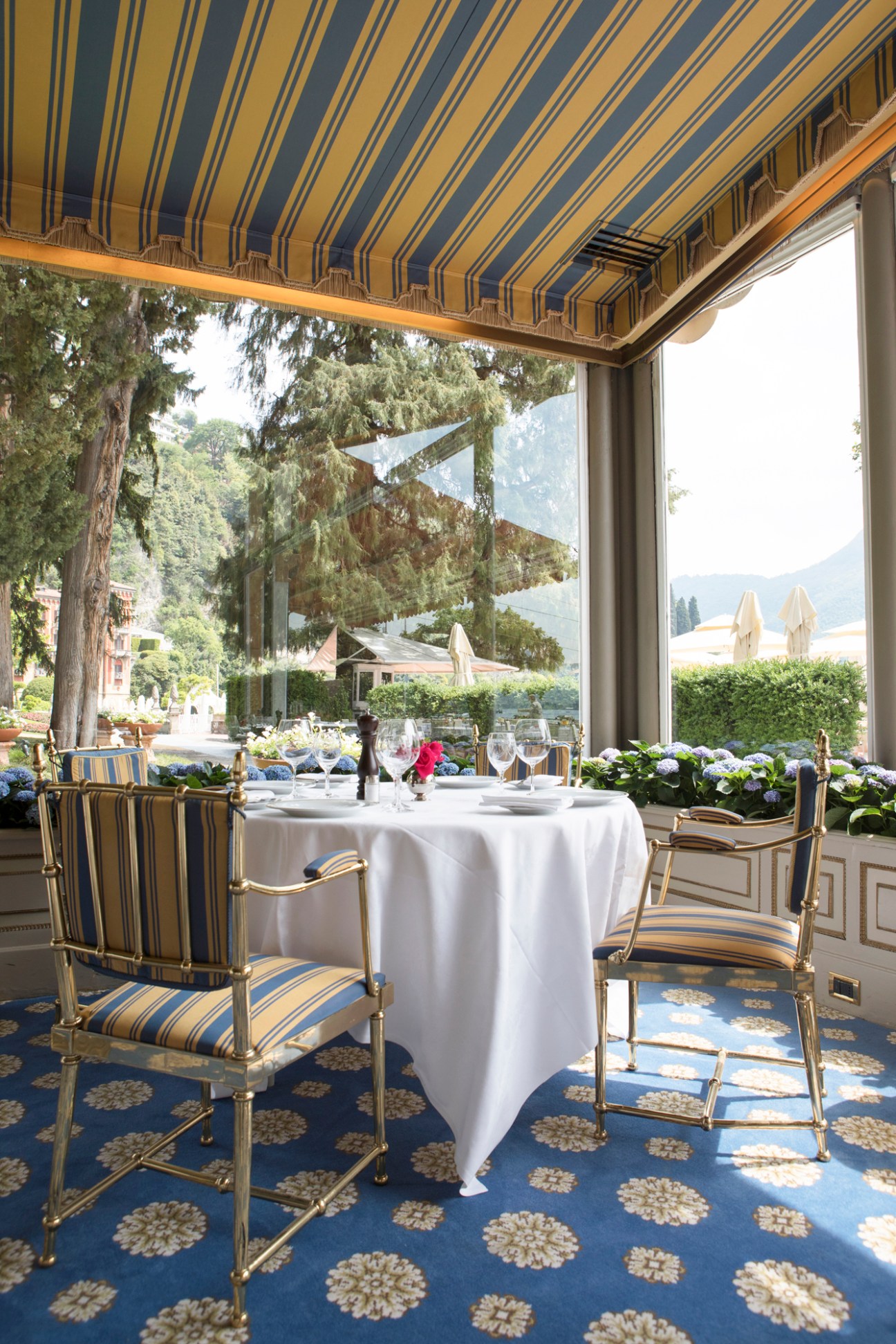 Elegant hotel dining terrace with striped awning, set table, ornate chairs and lake view through large windows