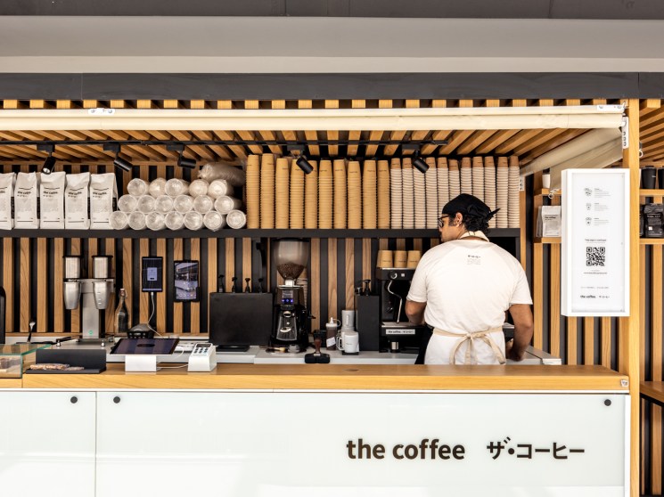 Barista in white apron working behind modern coffee shop counter with Japanese-inspired wood slat design and stacked cups