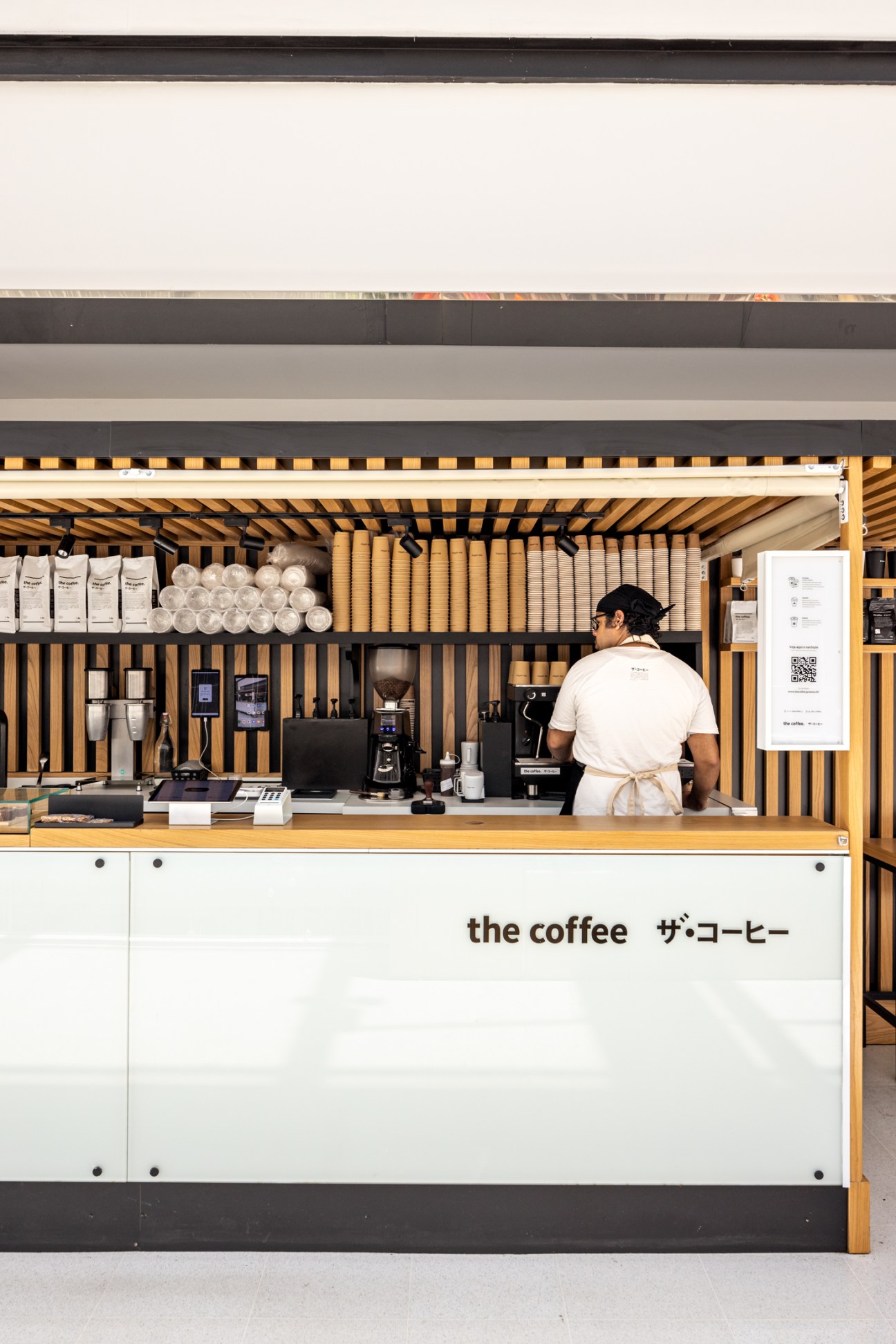 Barista in white apron working behind modern coffee shop counter with Japanese-inspired wood slat design and stacked cups