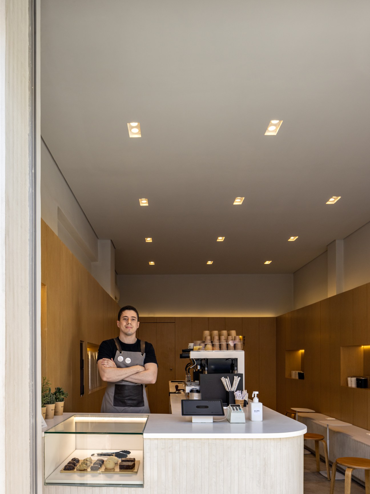 Barista in apron standing behind white counter with tablet ordering system and coffee machine in modern minimalist cafe