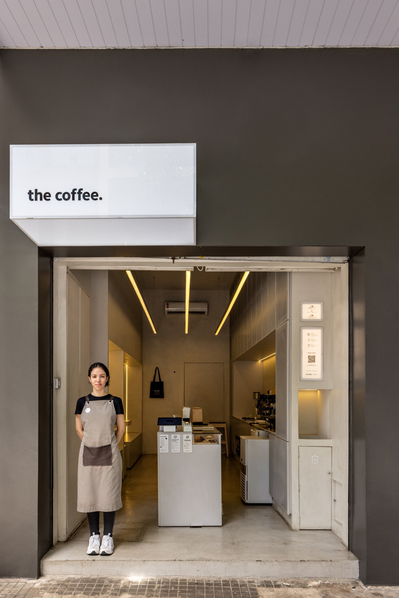 A female barista in an apron stands at the entrance of a small, minimalist coffee shop with modern lighting