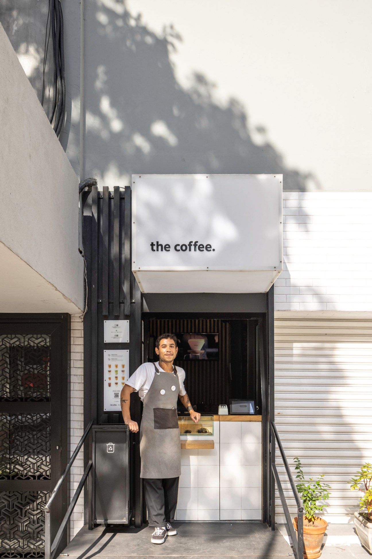 Smiling barista in apron standing behind counter at small The Coffee shop storefront