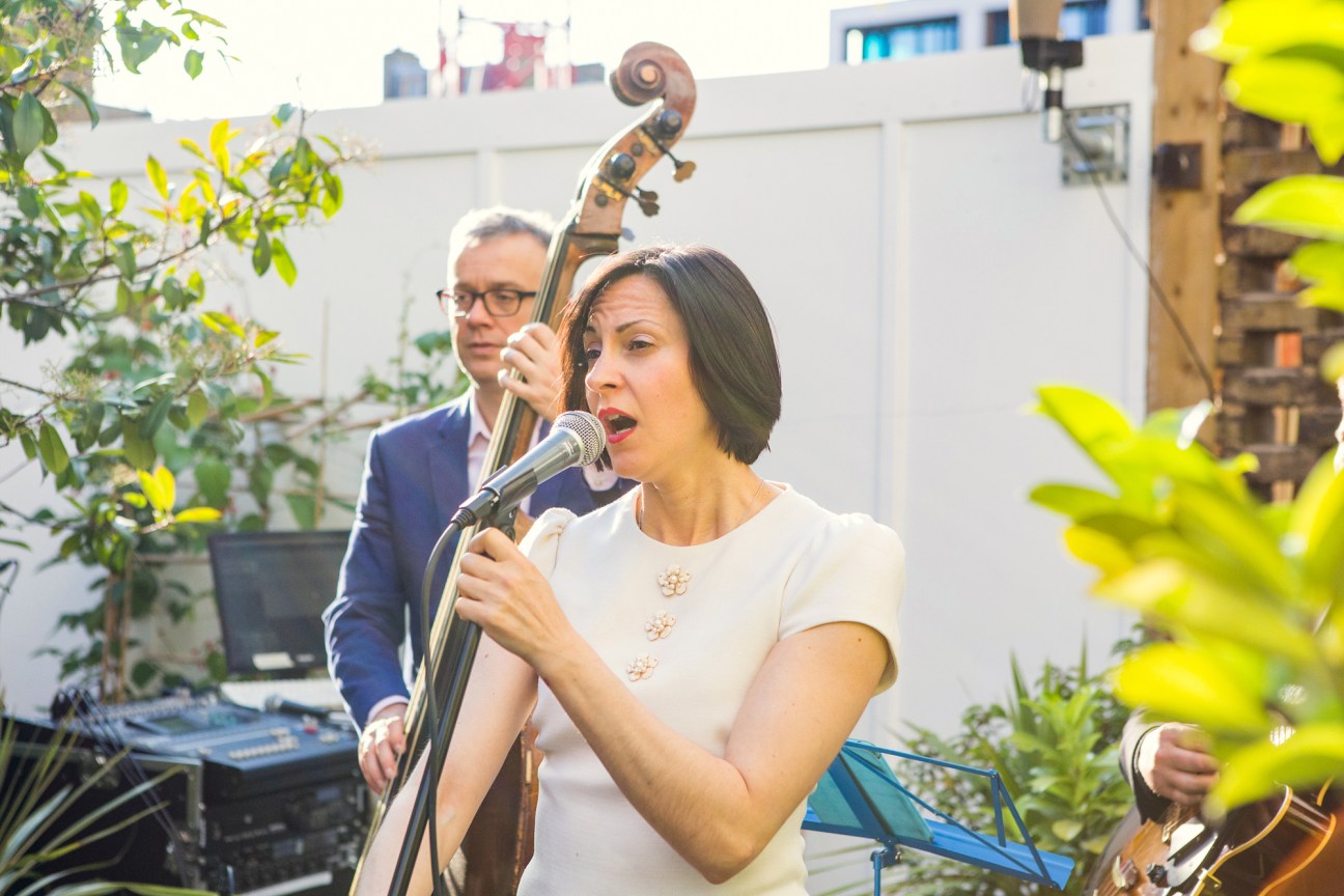 Woman in white top singing into microphone with man playing upright bass in outdoor courtyard setting