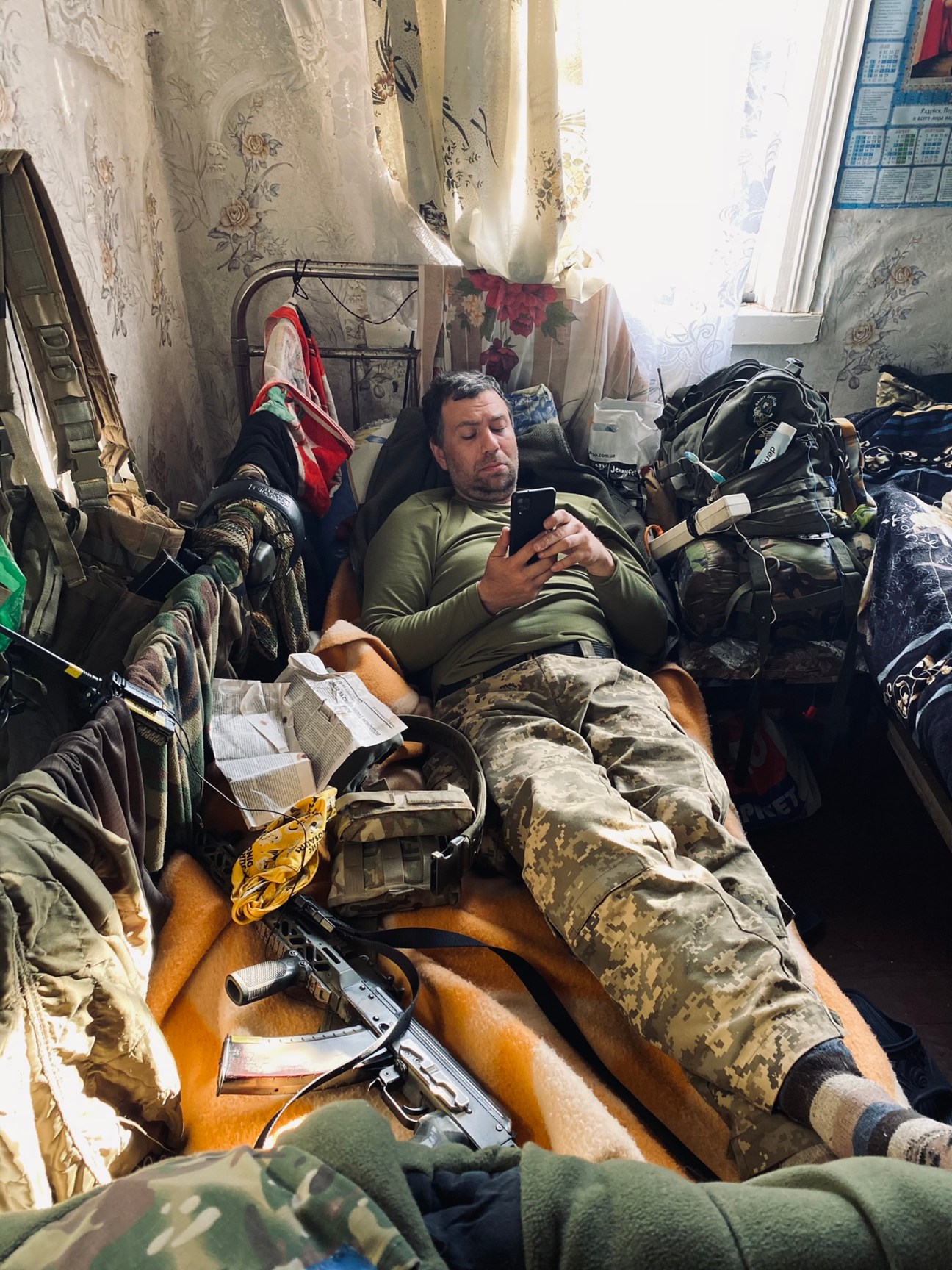 A soldier in camouflage uniform lying on a bed using a mobile phone in a cluttered room