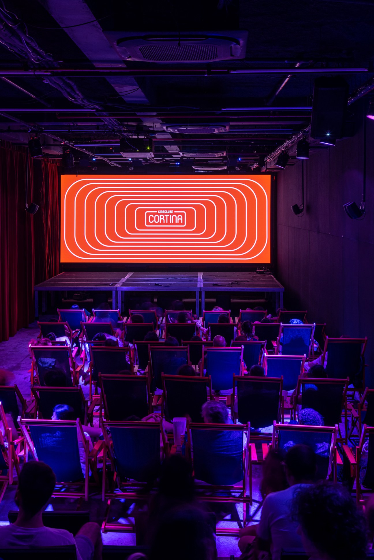 Cinema auditorium with red neon lighting, purple ambient lighting, and audience members seated watching a screen