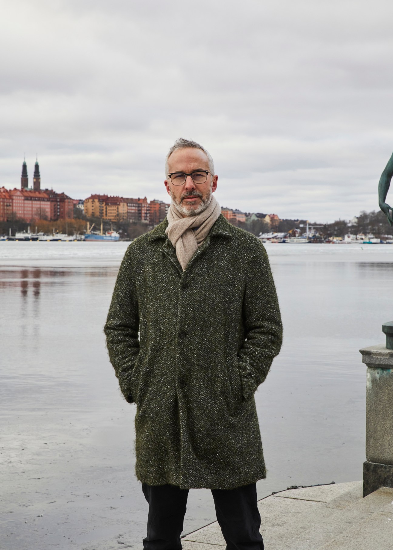 Dan Hill standing by waterfront wearing dark coat and scarf with Nordic cityscape in background