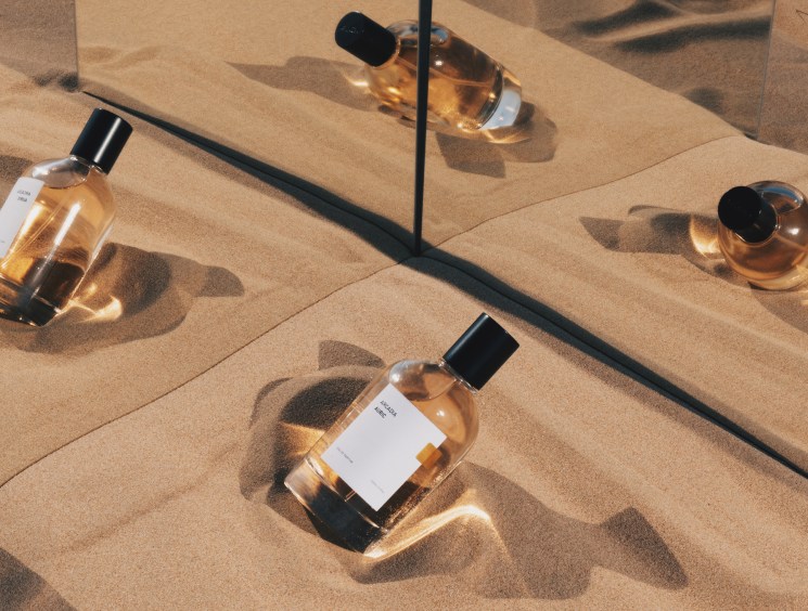Arcadia perfume bottles with golden liquid arranged on sand with dramatic shadows in sunlight.
