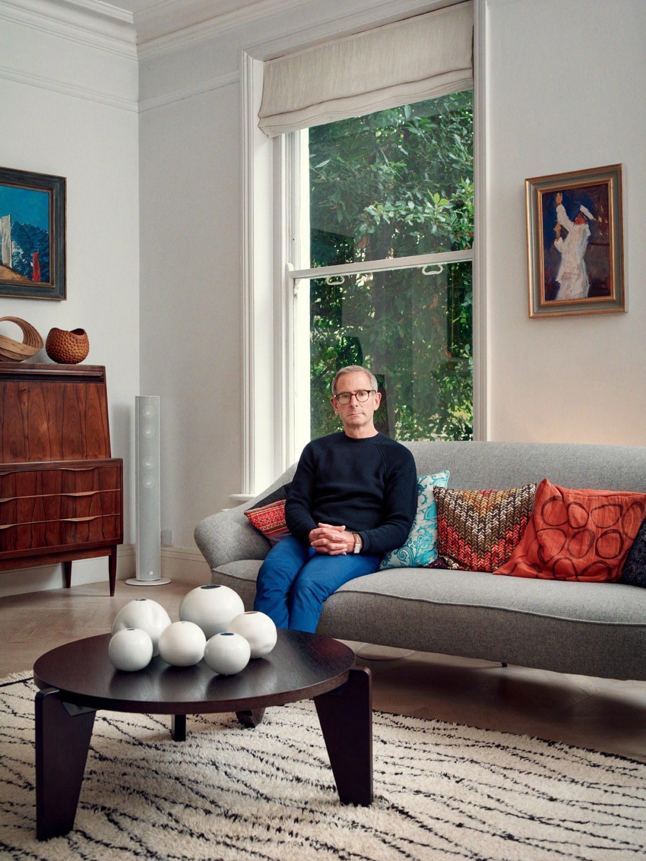 A man sitting on a gray sofa in a bright, modern living room with contemporary and mid-century furniture and artwork.
